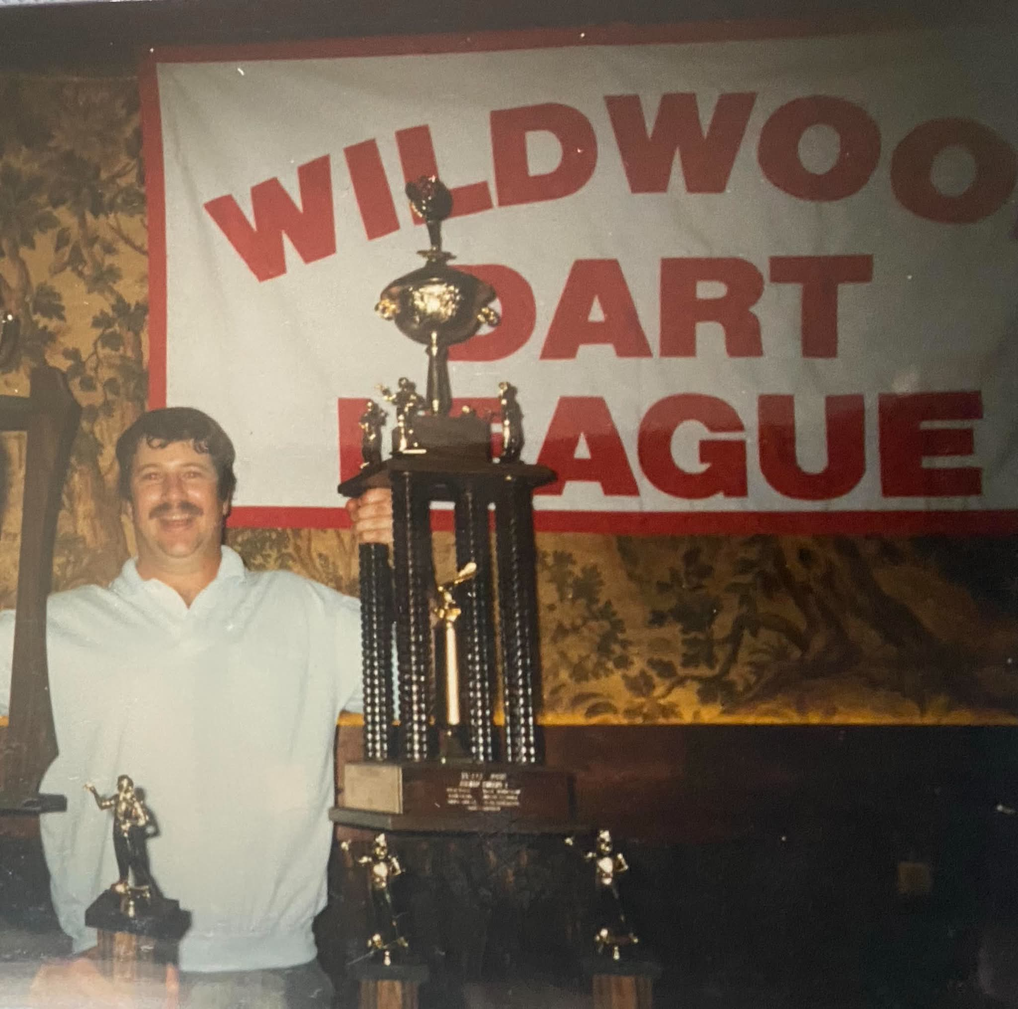 Don holding trophies in front of a Wildwood Dart League sign